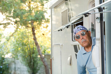 Uomo anziano con una bandana fantasia e occhiali da sole che si affaccia dal portellone del proprio camper, sullo sfondo un paesaggio naturale.