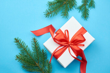 White gift box with red ribbon bow with fir tree branches on blue paper background