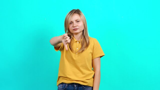 The young woman initially approved by raising her finger and finally disapproved. Beautiful Caucasian woman in yellow t-shirt and long blond hair standing on blue background. A fat teenager