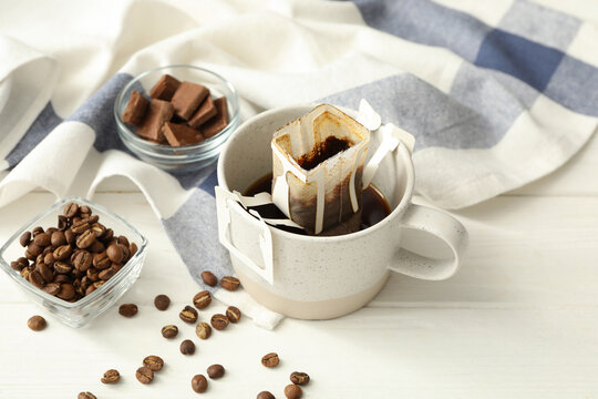 Cup with drip coffee bag and beans on white wooden table, closeup