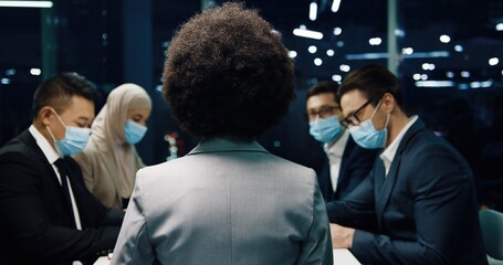 Rear of African American female boss speaking with team on meeting in modern office in evening. Mixed-races professional traders in medical masks discussing business issues. Conference concept