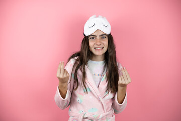 Pretty girl wearing pajamas and sleep mask over pink background doing money gesture with hands, asking for salary payment, millionaire business