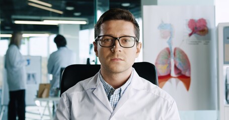 Close up portrait of handsome Caucasian male physician in glasses sitting in hospital office at workplace looking at camera and speaking on medical consultation. Healthcare concept. Doctor appointment