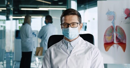 Fototapeta na wymiar Close up portrait of handsome young Caucasian adult male doctor wearing medical mask sitting in clinic at workplace looking at camera and smiling. Coronavirus pandemic. Hospital concept