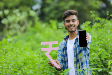 Young indian man holding indian rupees symbol in hand over nature background