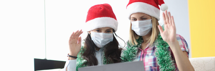 Two woman in protective mask and santa claus hat sit side by side and wave hello. Girlfriend talk...