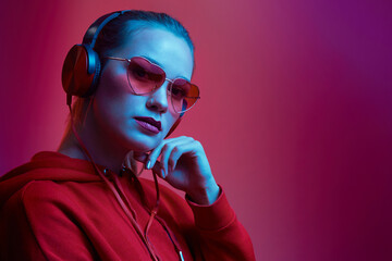 Fashion hipster woman wear stylish glasses and headphones listening to music over color neon background at studio.