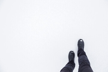 Young man in black leather boots walking on white snowy trail. Footwear for daily walking in winter season. Empty place for text on snow background. Point of view shot. Closeup. Top down view.