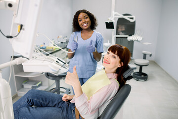 Healthy teeth and caries prevention concept. Young European woman at the dentist's chair during a dental examination and treatment, showing thumb up. Young African doctor dentist with dental tools