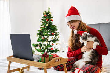 Happy young woman in santa hat sitting on bed with cat, dancing and looking at laptop. Online Christmas and new year celebration with pets. New reality. Online party and family congratulations