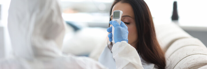 Doctor in protective suit measure patient temperature with non-contact thermometer. Portrait of sick woman on sofa.