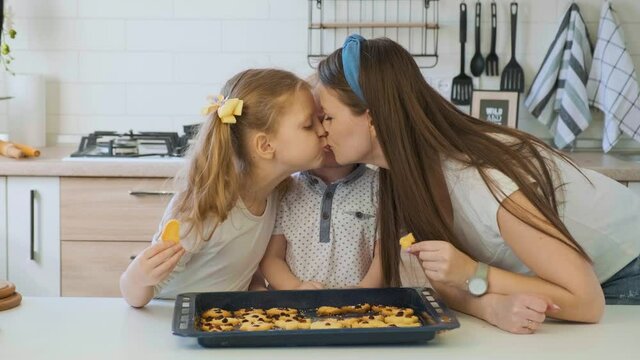 Beautiful young woman with two children taste prepared sweet homemade cookies and kissing with kids. Happy family together in the kitchen. Cooked delicious biscuits on the table.