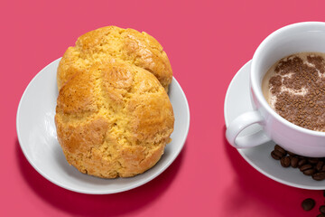 TWO COOKIES ON A WHITE PLATE AND A CUP OF COFFEE WITH MILK AND SOME COFFEE BEANS ON A PINK BACKGROUND