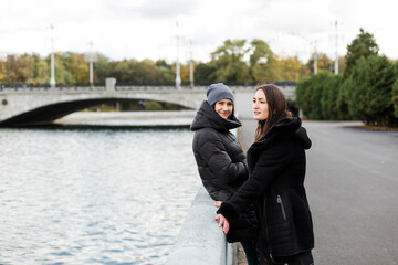 Two girls in warm clothes stand on the embankment by the water.