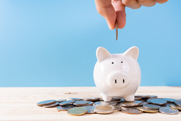 Hand putting money coin into piggy bank with a pile coins isolated blue background. Saving money...
