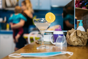 Home party in new normal, everyday life in lockdown, work life balance. Glass with cocktail on the kitchen table near laptop in home clutter and mother with baby on background