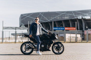A man in denim clothes sitting on a black sports motorcycle against the backdrop of a large modern building