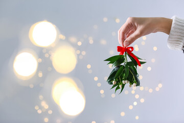 Woman holding mistletoe bunch with red bow against blurred festive lights, closeup and space for text. Traditional Christmas decor - obrazy, fototapety, plakaty