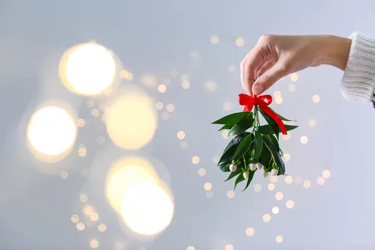 Woman holding mistletoe bunch with red bow against blurred festive lights, closeup and space for text. Traditional Christmas decor
