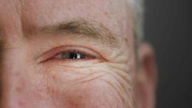 Extreme close up of happy and satisfied old man smiling with blue eye. Elderly mature male senior citizen with wrinkles laughing. Concept of health care, covid-19, retirement. Slow motion, macro, 4K.