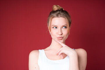 Cute blond woman with bun looks dreamy or thoughtful and looking to the side in white tank top isolated on red background.