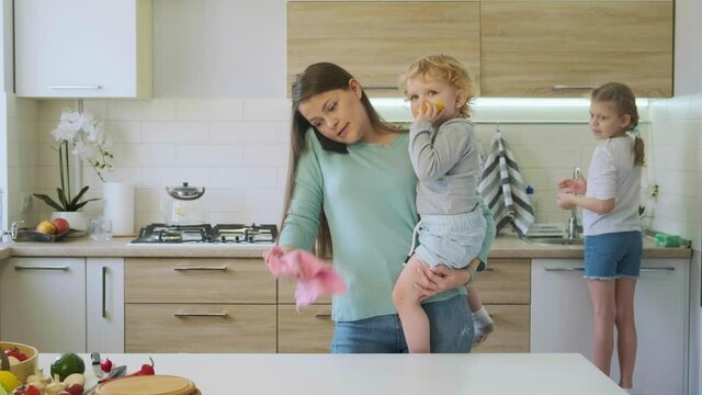 Close up busy young beautiful woman talking on the phone in the kitchen, hold little boy in hands and wipe the table. Girl washing plate on the background. Mother look after children and doing chores.