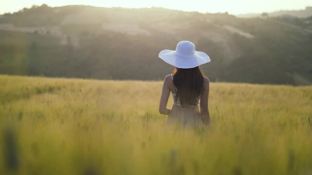Young woman relaxing in wheat field during beautiful warm sunset in summer. Cute brunette girl with hat enjoying romantic summertime atmosphere in nature. Italian countryside in Le Marche, Italy. 4K.