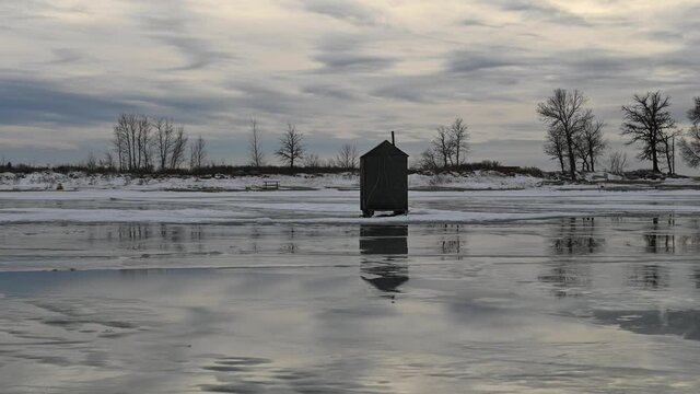 Time lapse of dramatic winter clouds moving through the sky and reflecting in water pooled on a frozen lake.  An ice fishing shack is in the foreground and trees in the mid-ground.
