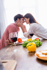 Smiling woman touching face of boyfriend near wine and vegetables on blurred foreground