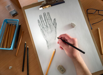 On the table are a drawing tablet with a sheet of paper, pencils, glasses and erasers. The human hand draws with a pencil on paper. Drawing training, lessons, hobbies.
