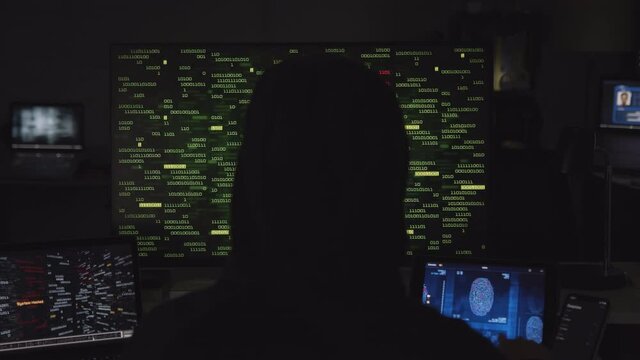 Hooded hacker working on computer. Dark room with devices playing several videos: internet security, program code security interface and fingerprint access. Technology. Cyber security. 