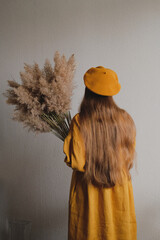 Woman in mustard yellow linen puff sleeve dress indoors. Girl holding a bouquet of high dry wild grass flowers.