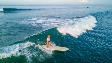 Drone shot of blonde surfer girl surfing on longboard at sunrise on Canggu beach, Bali - obrazy, fototapety, plakaty