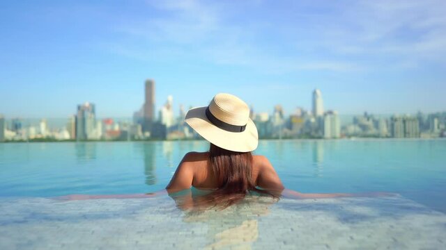Backside view of a Pretty young model wearing a swimming suit and hat and sitting inside infinity pool enjoying the city view, hotels rooftop