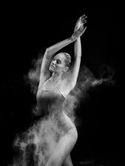 A beautiful slender ballet dancer girl wearing a bodysuit, sensually poses among the flying flour which covers her body, on a black background. Artistic, commercial, monochrome design.