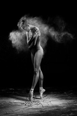 Fototapeta na wymiar A beautiful slender ballet dancer girl wearing a bodysuit and pointe shoes, posing dancing among the clouds of flying flour on a black background. Artistic, commercial, monochrome design
