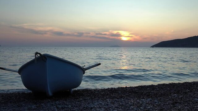 A lone row boat sits on a pebbly beach as the sun sets behind it and gentle waves roll in