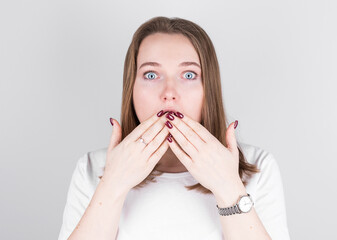 Young woman in a white T-shirt stands against a gray wall and covers her mouth with her hands in great surprise.