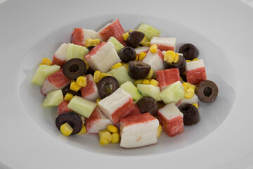 Spring salad. Closeup view of a colorful fresh salad served on a white bowl on the marble table. The salad contains sliced cucumber, crab sticks, corn grains and black olives.