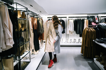 A beautiful woman takes off a beige coat from a hanger in a store and tries it on herself.