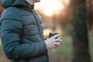 Fototapeta premium The guy holds a paper cup with coffee or tea. Have a cup of coffee. Disposable paper cup. Hot drink. Empty space. Man standing in the street holding a cup of coffee. Good morning.