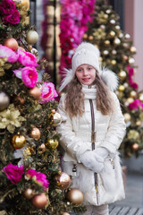 Portrait of a sweet smiling girl in a white knitted cap and white coat standing near decorated Christmas tree on a cold winter day outdoors