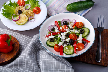 White plate with grilled zucchini with cherry tomatoes on lettuce and Greek salad on a napkin with a fork near vegetables.