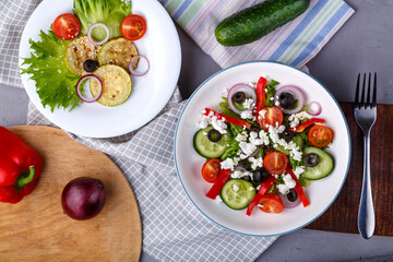 White plate with grilled zucchini with cherry tomatoes on lettuce and Greek salad on this table on a napkin with a fork near vegetables.