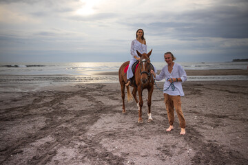 Horse riding on the beach. Woman on a brown horse. Man leading horse by its reins. Love to animals. Husband and wife spending time together. Bali, Indonesia