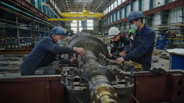 Workers discussing and working on a rotor of a steam turbine. The rotor is turned by steam impinging against attached vanes or blades on which it exerts a force. Turbine manufacturing plant.