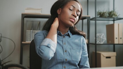 Young beautiful African-American woman in denim shirt works in office and sitting at table feels sharp pain in neck.
