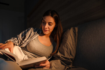 Charming focused woman reading book while lying in bed at home