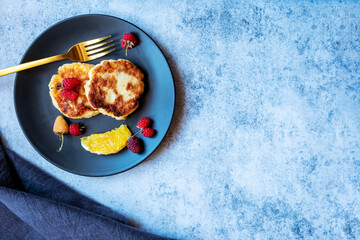 Cottage cheese pancakes, Russian syrniki, on a black plate with berries and orange. Healthy breakfast rich in calcium. With copy space.