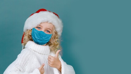 Merry Christmas on quarantine coronavirus pandemic. Little girl in Santa hat showing thumb up wearing medicine mask on blue color background. Child safety social distance